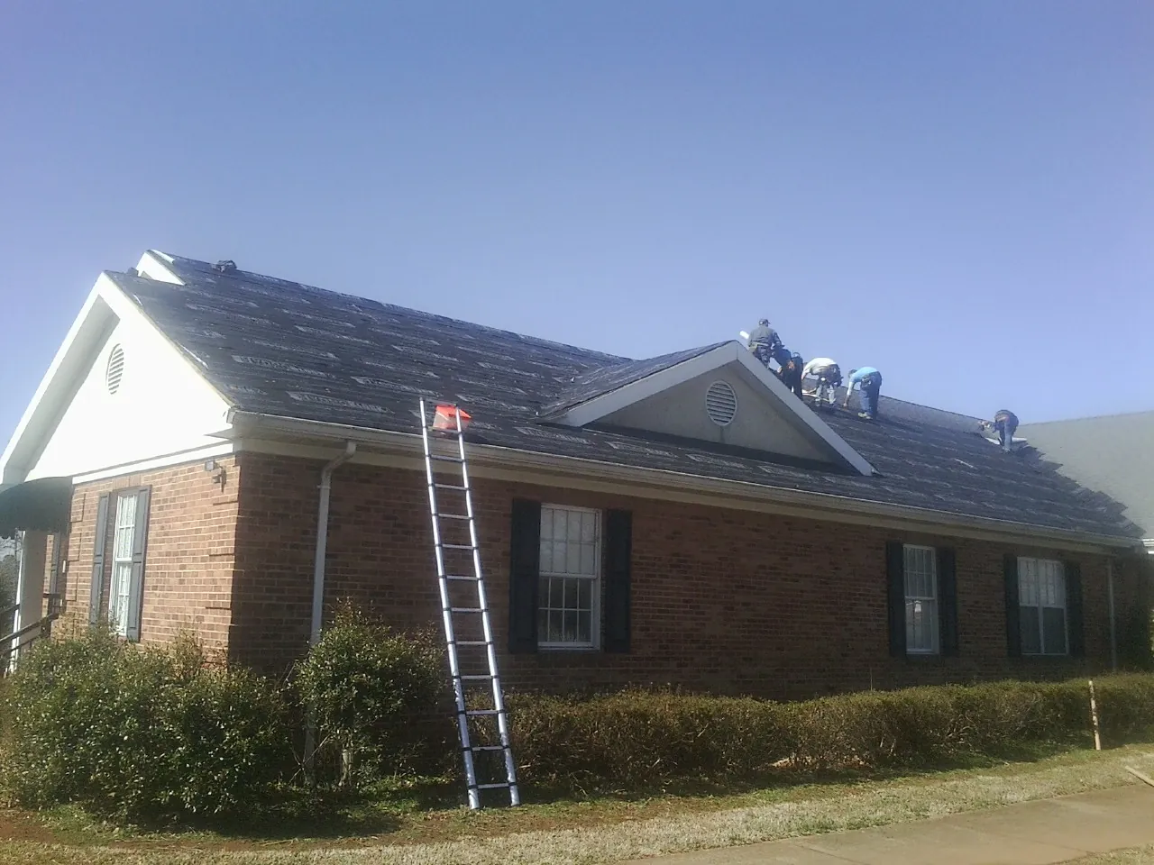 Crew tearing shingles off a brick ranch roof with a ladder set — Emergency Roof Repair work by our crew in Corinth